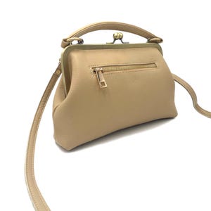 May include: A beige leather handbag featuring a structured frame with a brass-toned kiss-lock closure, a top handle, and a detachable shoulder strap. The front of the bag includes a horizontal zippered pocket with a matching brass-toned pull tab.
