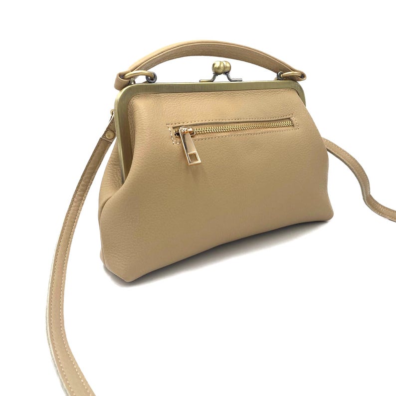 May include: A beige leather handbag featuring a structured frame with a brass-toned kiss-lock closure, a top handle, and a detachable shoulder strap. The front of the bag includes a horizontal zippered pocket with a matching brass-toned pull tab.