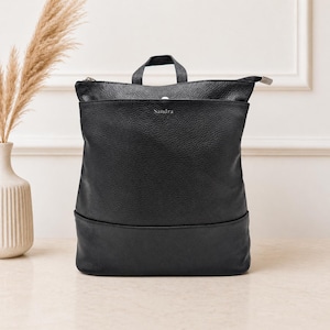 May include: Black leather backpack with a top handle and a zippered closure. The backpack has a textured surface and the name "Sandra" is embossed on the front. The bag is sitting on a light-colored surface.