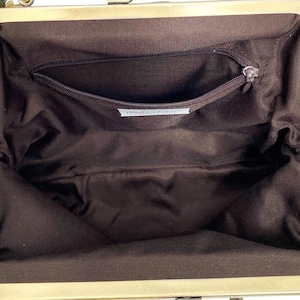 May include: The interior of an open handbag featuring a dark brown fabric lining. A brass-toned metal frame surrounds the opening. A single zippered pocket is located on the back wall of the interior, with a small white rectangular label attached below.