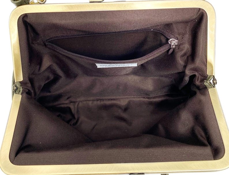 May include: The interior of an open handbag featuring a dark brown fabric lining. A brass-toned metal frame surrounds the opening. A single zippered pocket is located on the back wall of the interior, with a small white rectangular label attached below.