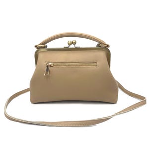 May include: A beige leather handbag featuring a structured frame with a brass-toned kiss-lock closure and a top handle. The front of the bag includes a horizontal zippered pocket with a gold-toned pull and a long, thin, adjustable shoulder strap.