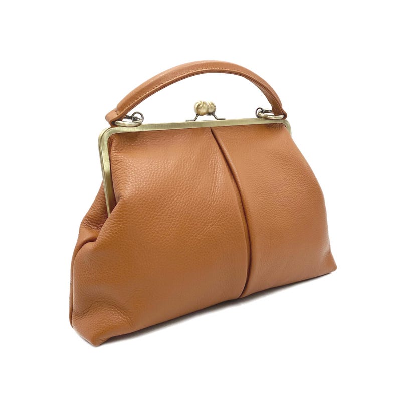 Puede incluir: Un bolso de mano de cuero color canela con superficie texturizada, que presenta un asa superior curva y un cierre de marco de metal en tono lat&oacute;n con un broche de doble bola. El bolso tiene una forma estructurada con una costura central.