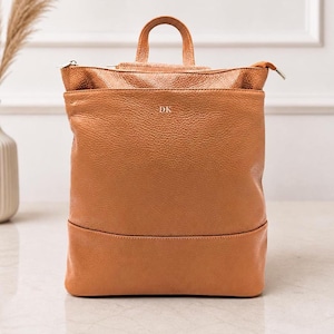May include: A tan leather backpack with a top handle and a zippered closure. The backpack has a rectangular shape with a textured surface and the initials "DK" are embossed on the front. The bag is sitting on a light-colored surface.
