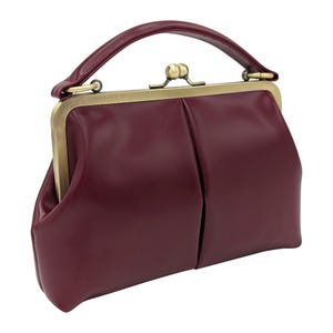 K&ouml;nnte beinhalten: Burgunderfarbene Handtasche mit oberem Griff und Schnallenverschluss. Die Tasche hat einen Vintage-Stil mit strukturierter Form und goldfarbenem Metallrahmen. Der Griff ist gebogen und die Tasche scheint aus Leder oder einem &auml;hnlichen Material zu sein.