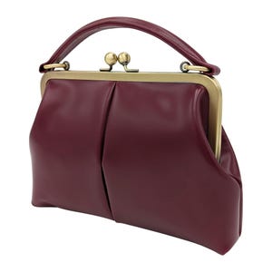 K&ouml;nnte beinhalten: Eine weinrote Handtasche mit einem oberen Griff und einem Schnallenverschluss. Die Tasche hat eine strukturierte Form mit einem plissierten Design auf der Vorderseite. Der Verschluss und die Griffbefestigungen sind goldfarben.