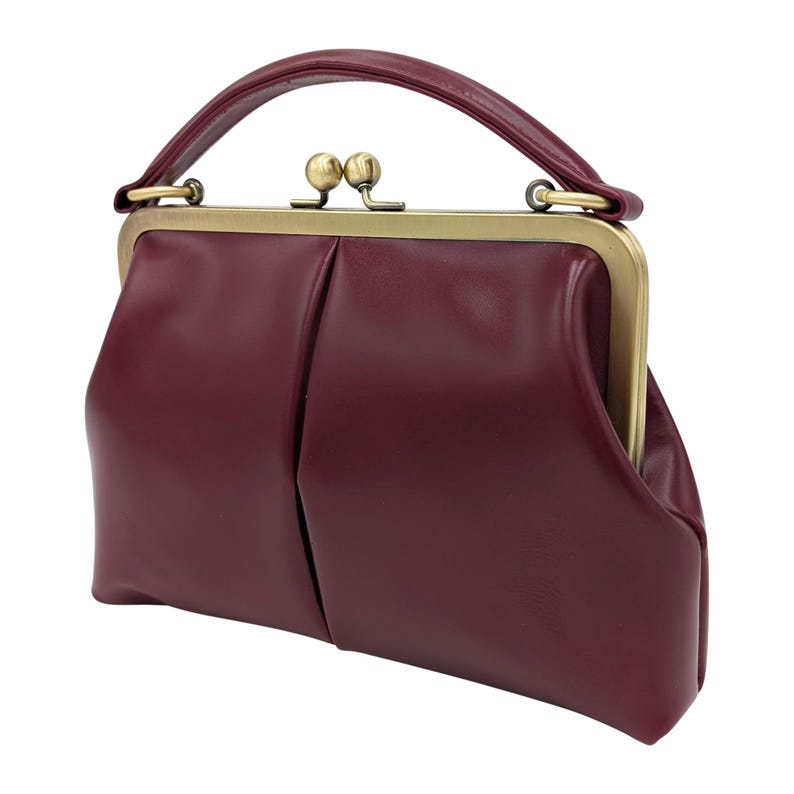 K&ouml;nnte beinhalten: Eine weinrote Handtasche mit einem oberen Griff und einem Schnallenverschluss. Die Tasche hat eine strukturierte Form mit einem plissierten Design auf der Vorderseite. Der Verschluss und die Griffbefestigungen sind goldfarben.