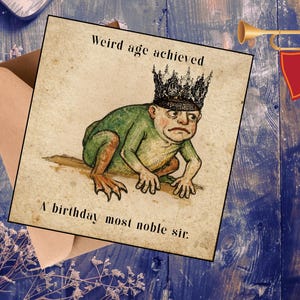 May include: A birthday card featuring a frog-like figure wearing a crown, with the text "Weird age achieved" and "A birthday most noble sir." The card is on a wooden surface with a trumpet and envelope.