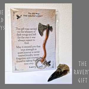 May include: A metal axe keyring with an orange handle and silver blade featuring intricate engravings, attached to a card titled The Old Ways The Raven's Gift, which includes text about strength and loyalty, alongside a small raven skull replica.