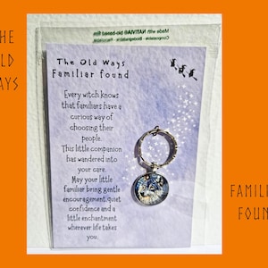 May include: A silver-toned circular keyring featuring a wolf portrait under a glass dome, attached to a light purple card with the text The Old Ways Familiar found and a printed message about familiars, all enclosed in a clear plastic sleeve.