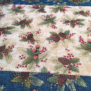 May include: A beige fabric with a repeating pattern of green leaves, red berries, and brown pine cones. The fabric has a blue border with a similar pattern.