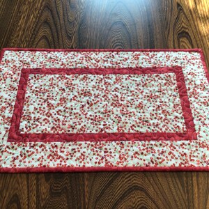 May include: Rectangular quilted placemat with a white background covered in small red cherry patterns. The placemat has a red border and a central rectangular design with the same cherry pattern. The quilting adds texture.