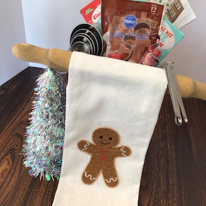 May include: A white kitchen towel with a brown gingerbread man applique is the focal point of this image. A rolling pin, measuring cups, and gingerbread house kit are also visible. A small, silver tinsel tree adds a festive touch.