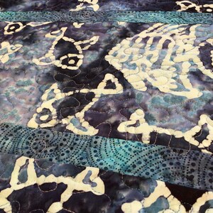 May include: A close-up of a quilt with a blue and purple batik pattern. The quilt is made of fabric with a white design of fish and other shapes. The quilt is quilted with a dark thread.