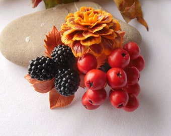 May include: A decorative brooch featuring a cluster of three textured black blackberries, a vibrant orange and yellow marigold flower, and a bunch of bright red berries, all accented with small brown leaves arranged in a naturalistic composition.