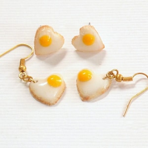 May include: A pair of heart-shaped earrings with a fried egg design. The earrings are made of white and yellow polymer clay and have gold hooks.