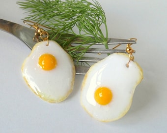 May include: A pair of gold earrings shaped like fried eggs with yellow yolks. The earrings are hanging from a fork.