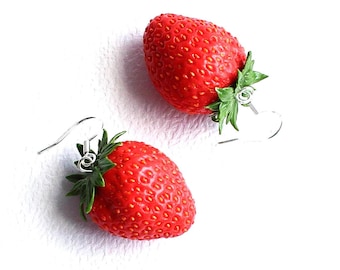 May include: A pair of strawberry earrings. Each earring features a bright red, textured strawberry with green leaves and a silver-colored hook. The earrings are set against a white background.