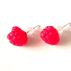 May include: A pair of bright red raspberry stud earrings. Each earring features a detailed, textured raspberry design with a silver post and a clear earring back. The earrings are set against a white background.