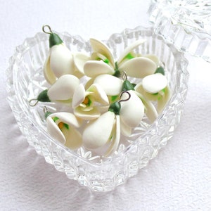 May include: A clear, heart-shaped glass trinket box with a lid, filled with small white and green flower charms. The charms have a delicate, handcrafted appearance. The box is open, revealing the contents, and sits on a white surface.
