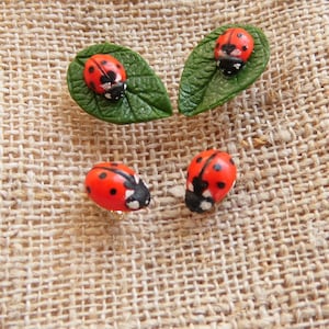 May include: Close-up of four ladybug-themed decorative items. Two ladybugs rest on green leaf-shaped bases, while two others are positioned separately. The ladybugs are red with black spots and black heads. The background is a textured, tan fabric.