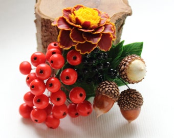 May include: A decorative floral arrangement featuring a deep red and yellow marigold, a cluster of bright red berries, small black berries, green leaves, and three brown acorns with textured caps, all gathered together in a compact, detailed display.