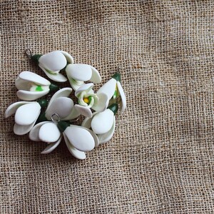 May include: A cluster of white, handcrafted snowdrop flower pendants with green and yellow accents. The pendants are arranged on a textured, tan burlap background, showcasing the delicate floral design. Each pendant has a small metal loop.
