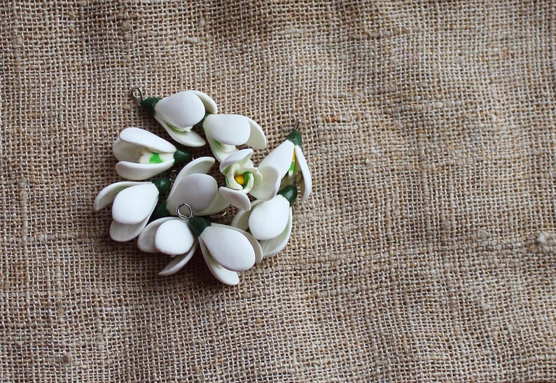 May include: A cluster of white, handcrafted snowdrop flower pendants with green and yellow accents. The pendants are arranged on a textured, tan burlap background, showcasing the delicate floral design. Each pendant has a small metal loop.