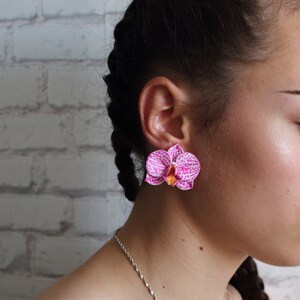 May include: A pair of pink orchid earrings with white and yellow accents. The earrings are worn by a person with dark hair.