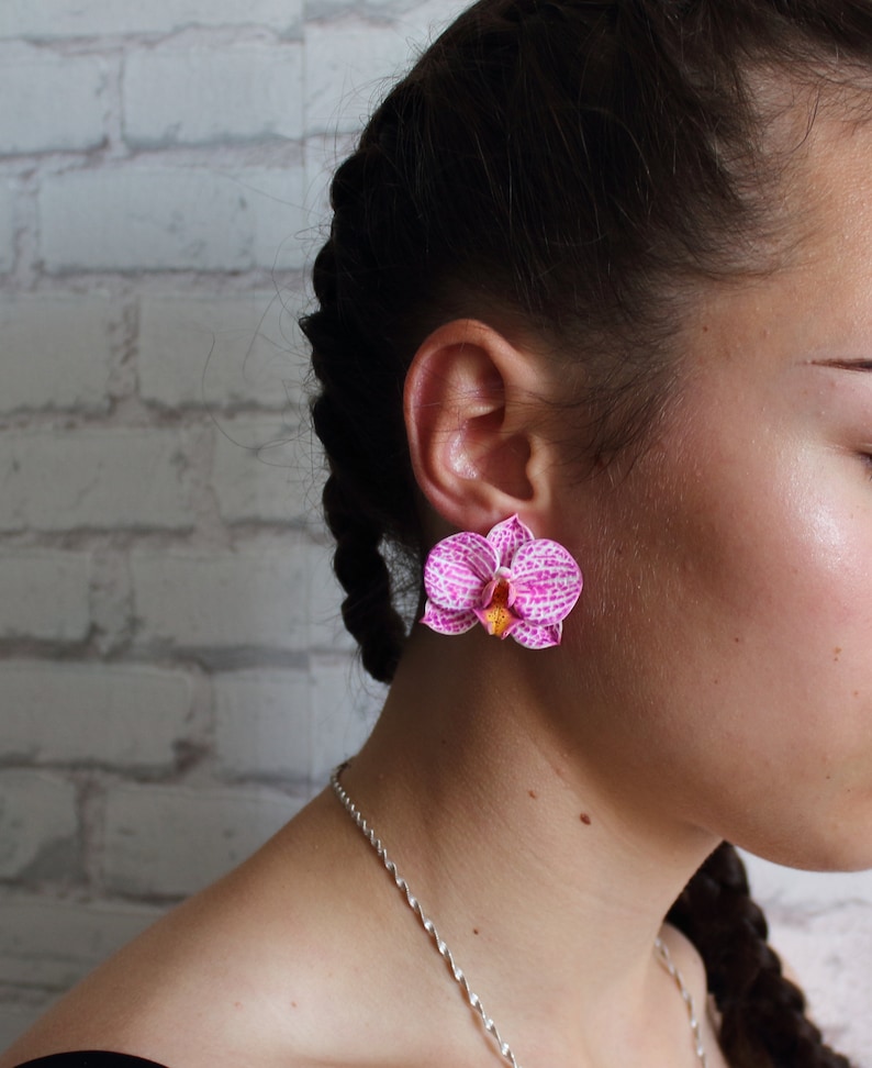 May include: A pair of pink orchid earrings with white and yellow accents. The earrings are worn by a person with dark hair.
