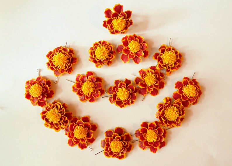 A collection of 15 orange and yellow marigold flowers arranged in a heart shape on a white background. Handmade polymer clay marigold bead flower.