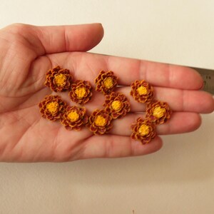 May include: A collection of ten small, brown and yellow felt flower embellishments. The flowers are arranged in a hand.