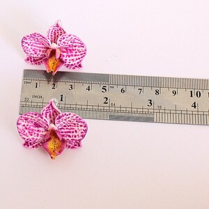May include: Two pink and white orchid flower embellishments with yellow centres, measured against a ruler. The flowers are approximately 1 1/2 inches wide.