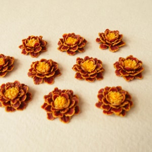 May include: Ten small, artificial marigold flowers with burgundy petals and yellow centers. The flowers are arranged in a symmetrical pattern on a white background.