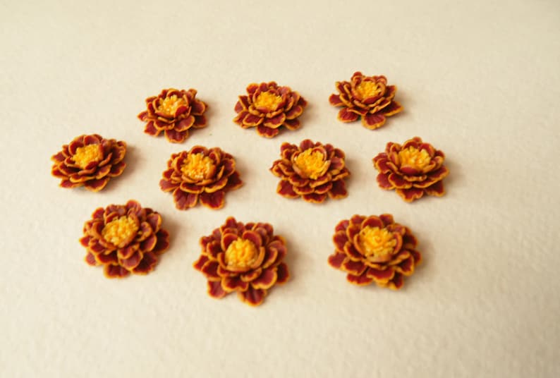 May include: Ten small, artificial marigold flowers with burgundy petals and yellow centers. The flowers are arranged in a symmetrical pattern on a white background.