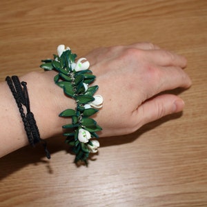 May include: A green and white floral bracelet with a silver chain is worn on a wrist. The bracelet features green leaves and white flower buds. A black woven bracelet is also on the wrist.