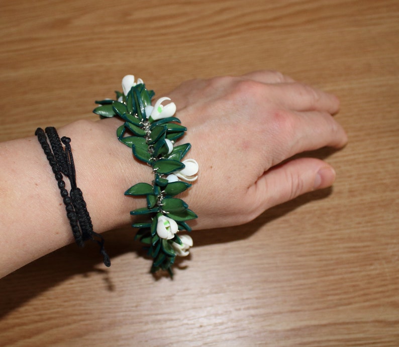 May include: A green and white floral bracelet with a silver chain is worn on a wrist. The bracelet features green leaves and white flower buds. A black woven bracelet is also on the wrist.