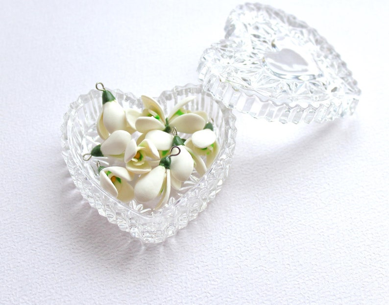 May include: A clear, heart-shaped glass trinket box with a lid, filled with small white and green flower charms. The charms have a delicate, handcrafted appearance. The box is open, revealing the contents, and sits on a white surface.