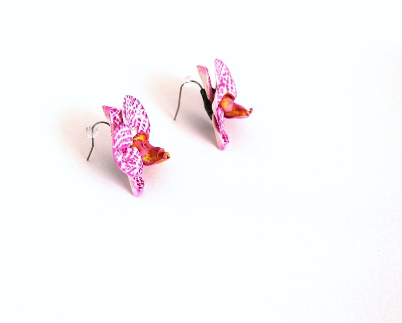 May include: A pair of pink and white orchid earrings with silver hooks. The orchids have a spotted pattern and a yellow centre.