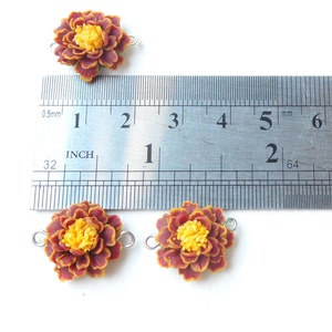 May include: Four brown and yellow marigold flower charms with silver jump rings. The charms are approximately 1 inch in diameter. A ruler is shown for scale.