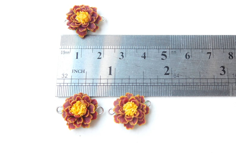 May include: Four brown and yellow marigold flower charms with silver jump rings. The charms are approximately 1 inch in diameter. A ruler is shown for scale.