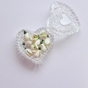 May include: A clear, heart-shaped glass trinket box with a lid, filled with small white and green flower buds. The lid features a white heart shape. The box is open, revealing the contents.