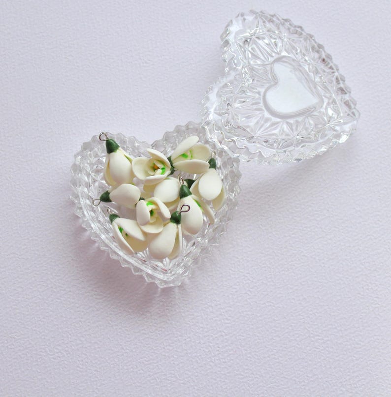 May include: A clear, heart-shaped glass trinket box with a lid, filled with small white and green flower buds. The lid features a white heart shape. The box is open, revealing the contents.