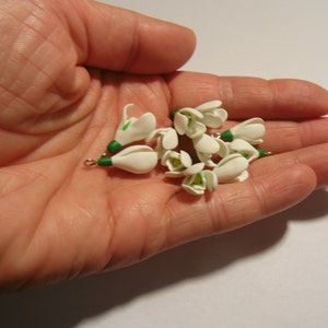 May include: A collection of small, white snowdrop flower charms with green accents, held in a hand. Each charm has a small metal loop. The flowers are clustered together, showcasing their delicate design and detail.