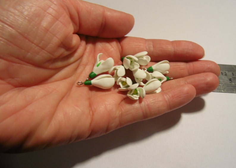 May include: A collection of small, white snowdrop flower charms with green accents, held in a hand. Each charm has a small metal loop. The flowers are clustered together, showcasing their delicate design and detail.