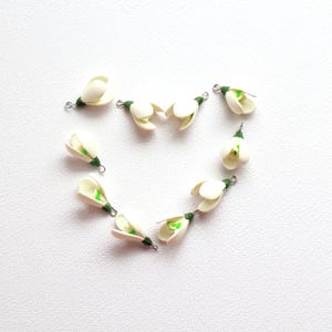 May include: A collection of small, white flower charms with green accents and silver-tone loops. The charms are arranged in a heart shape against a white background. These delicate floral charms are suitable for jewelry making or craft projects.