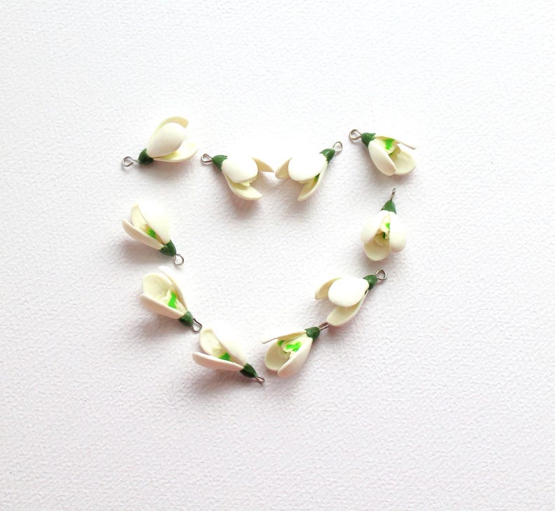 May include: A collection of small, white flower charms with green accents and silver-tone loops. The charms are arranged in a heart shape against a white background. These delicate floral charms are suitable for jewelry making or craft projects.