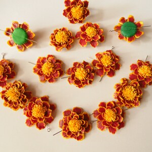 May include: A set of 14 orange and yellow marigold flower hair clips with green centers. The clips are arranged in a circle on a white surface.