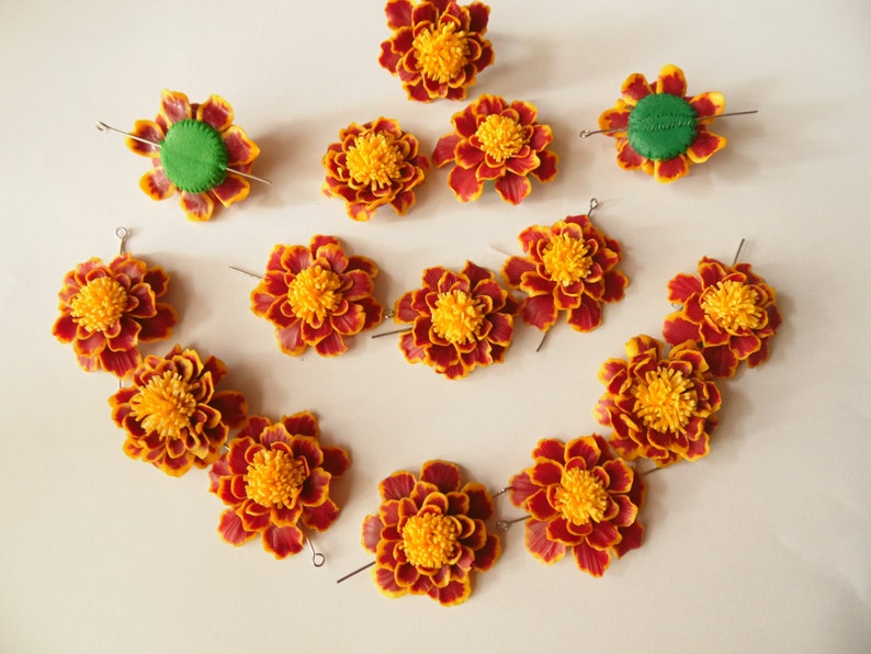 May include: A set of 14 orange and yellow marigold flower hair clips with green centers. The clips are arranged in a circle on a white surface.