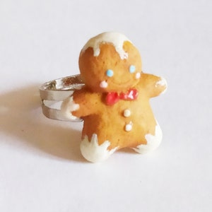 May include: A gingerbread man ring with a silver band. The gingerbread man is brown with white icing on the head and feet, blue eyes, a red bow tie, and white buttons. The ring is set against a white background.